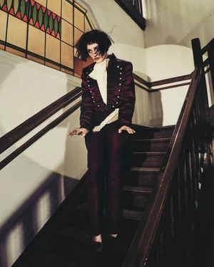 Model in a burgundy velvet jacket standing on dark wooden indoor stairs near a stained glass window