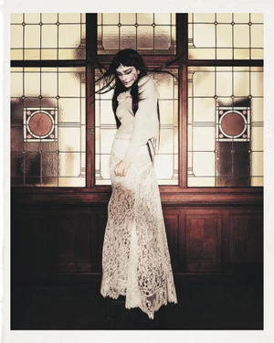Model with long dark hair wearing a sheer white lace dress standing in front of large stained glass windows.