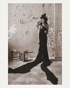 Model in a black one-shoulder dress with a long train standing near an overturned chair against a distressed peeling wall.