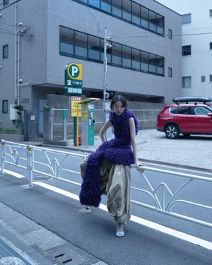 Model wearing a textured purple top and loose beige trousers leaning against a white metal street railing