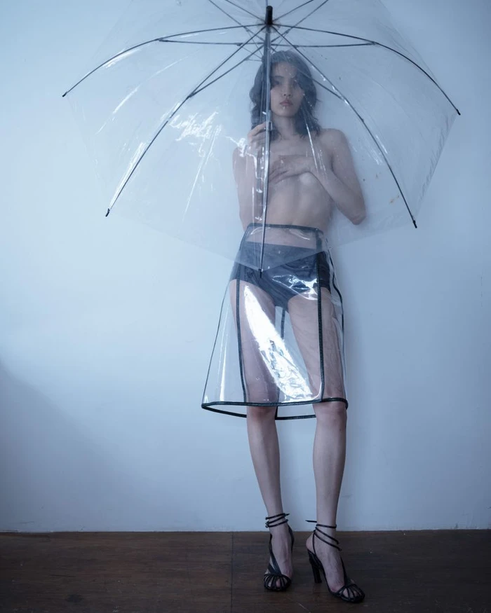 Model holding a clear umbrella wearing a transparent skirt and dark heels against a neutral background
