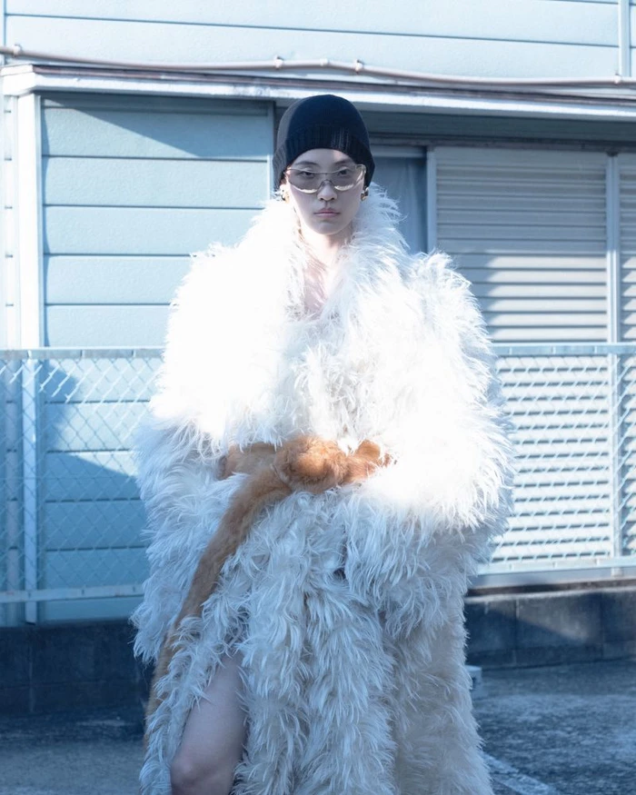 Model wearing a white shaggy textured coat, black beanie, and sunglasses standing outside a building exterior.