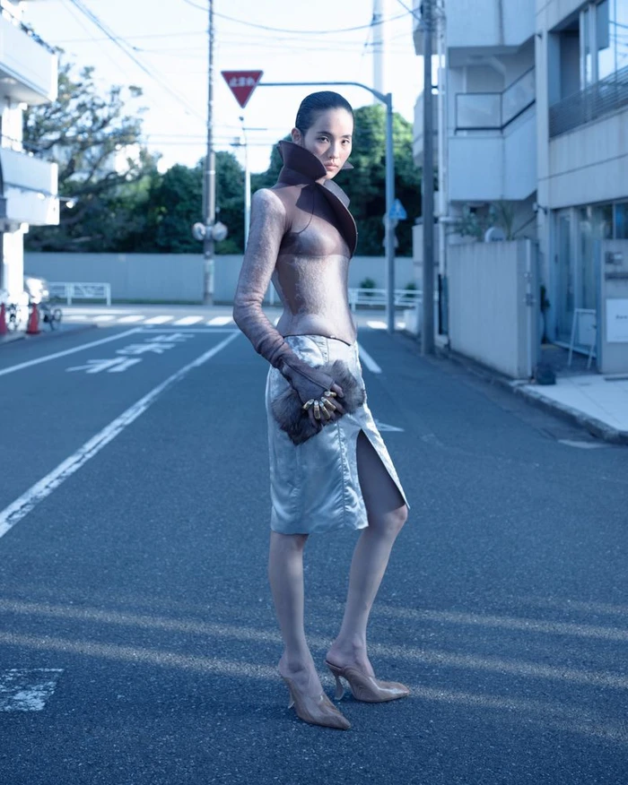 Model standing on a city street wearing a structured high-collar top and a side-slit skirt with clear heels.