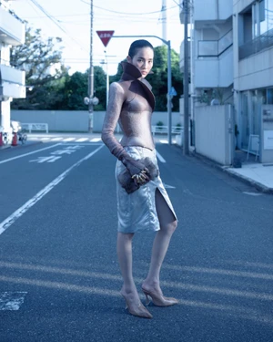 Model standing on a city street wearing a structured high-collar top and a side-slit skirt with clear heels.