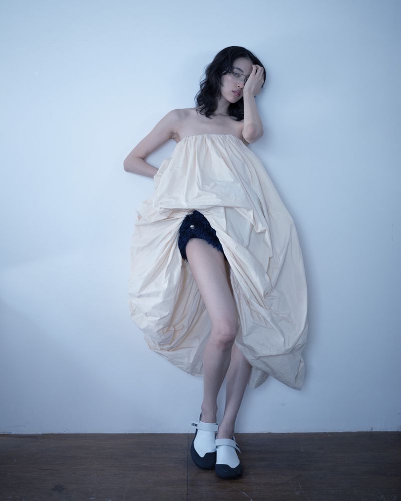 Model Song Ah Woo wearing voluminous cream dress and white shoes against a plain wall for Exhibition Magazine