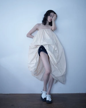 Model Song Ah Woo wearing voluminous cream dress and white shoes against a plain wall for Exhibition Magazine