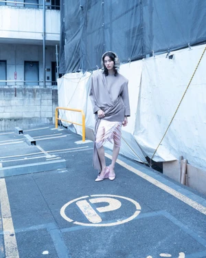 Model standing in an asphalt parking lot with a white construction barrier in the background for Exhibition Magazine.