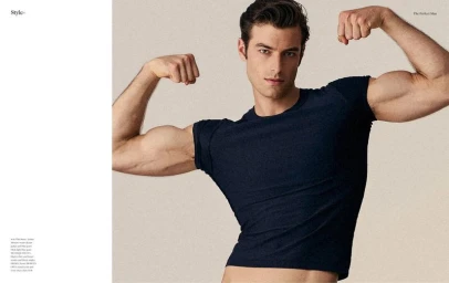 A dark-haired male model flexing his biceps in a dark blue t-shirt against a neutral studio background for The Perfect Man Magazine.