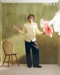 Man holding scissors and a large artificial pink flower in an olive green room with a wooden chair.