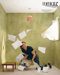 Man kneeling on a floor holding a typewriter as sheets of paper fly through the air in a green-walled room.