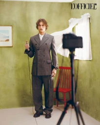 Man wearing a grey double breasted suit holding a camera cable in an olive green room with a red chair