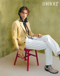 Male model seated on a red wooden chair wearing a beige blazer, white trousers, and patterned boots against an olive green backdrop