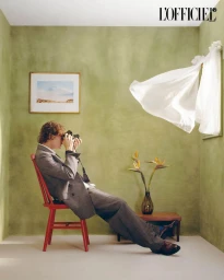 Photographer in a grey suit seated on a red chair in a green room holding a camera to his eye