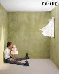 Man seated on the floor in a green textured room holding a multi-tiered strawberry cake as white curtain blows from a wall opening