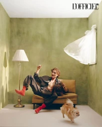 Man seated on a sofa with two dogs in a green room with a floor lamp and a floating white garment on the wall.