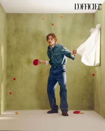 Young man in denim shirt and pants holding a ping-pong paddle in a green room with floating balls and a blowing curtain