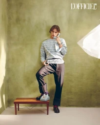 Young man in striped shirt holding a telephone receiver while standing with one foot on a small wooden bench against a textured green wall.