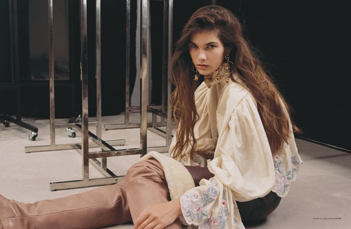 Model seated on floor wearing ruffled ivory blouse and tall leather boots against studio backdrop