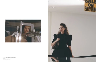 Model wearing a black dress with structured shoulders and holding a handbag in a retail store setting.