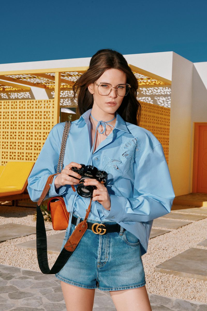 Young woman poses outdoors in a blue shirt, denim shorts, and Gucci belt, holding a camera, with a vintage aesthetic and bright blue sky.