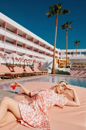 Woman in floral dress lounges by pool at pink hotel with cocktail, palm trees, and blue sky, enjoying vacation on sunny day.