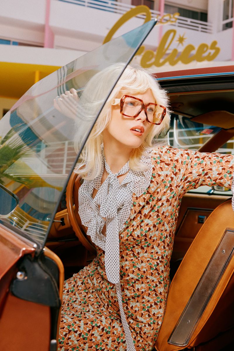 Stylish woman in patterned dress and oversized glasses exits vintage car in front of the Felices building with palm trees.