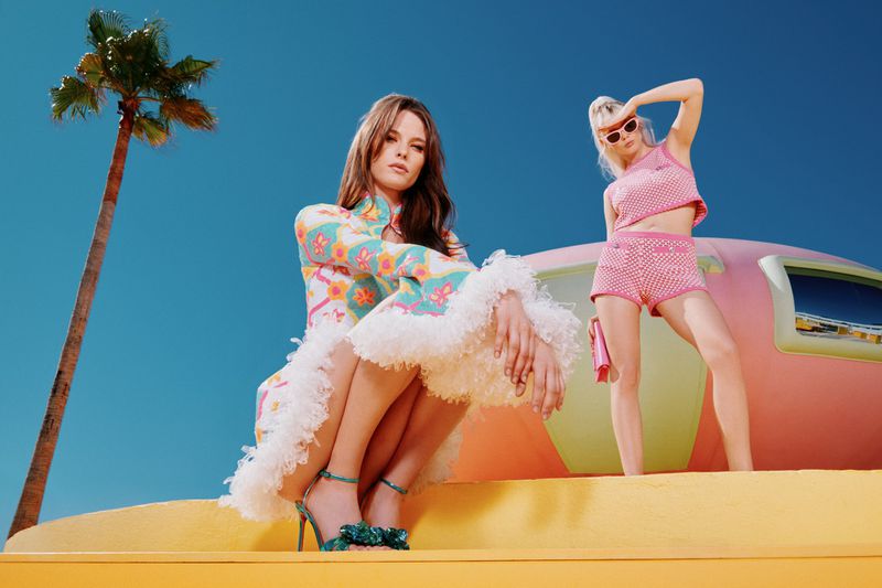 Two fashion models pose on a yellow and pink structure under a blue sky near a palm tree wearing vibrant, retro-inspired clothing.
