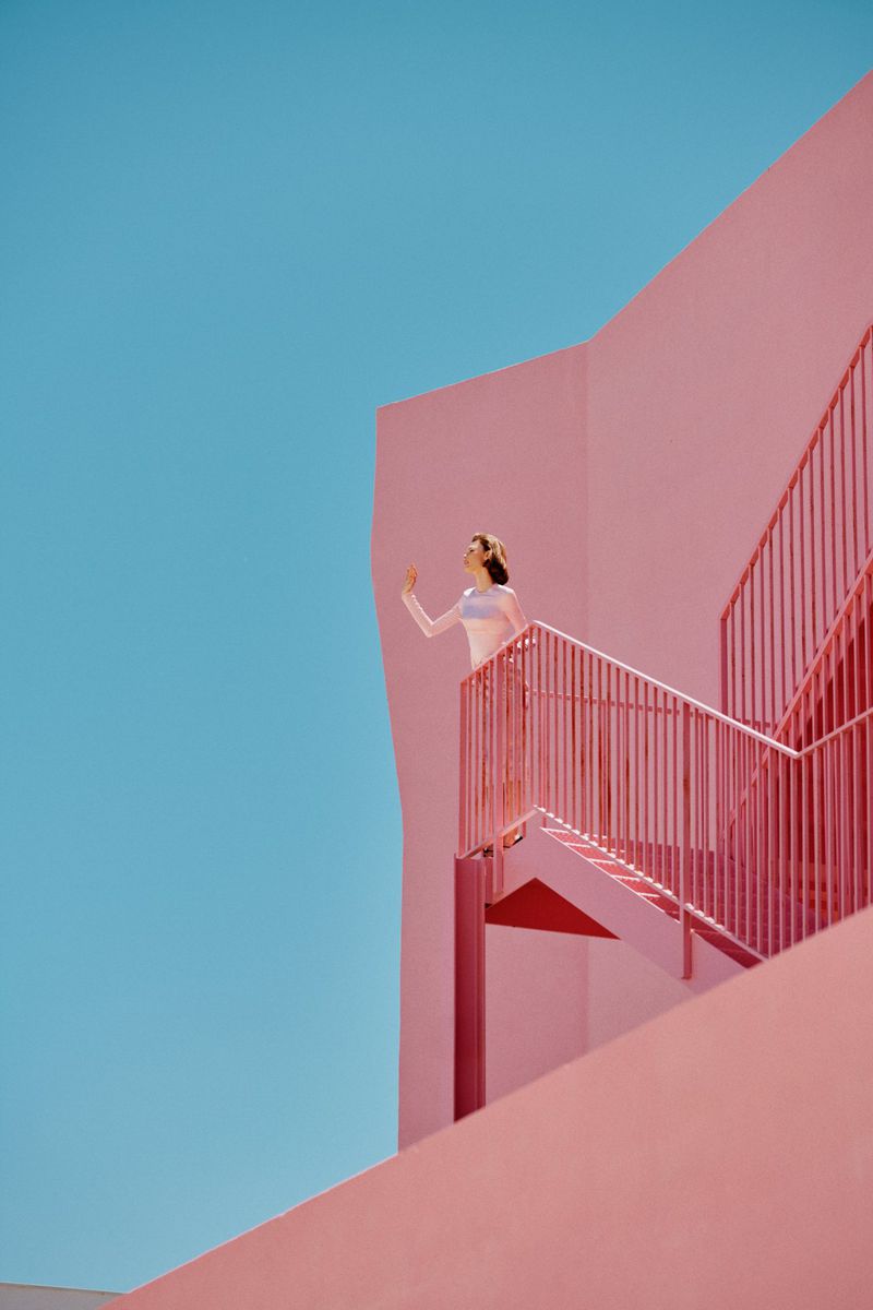 Woman standing on a pink stairway against a clear blue sky, wearing light clothing in a bright outdoor setting with architectural elements.