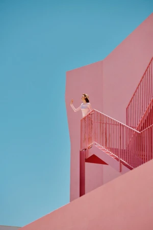 Woman standing on a pink stairway against a clear blue sky, wearing light clothing in a bright outdoor setting with architectural elements.