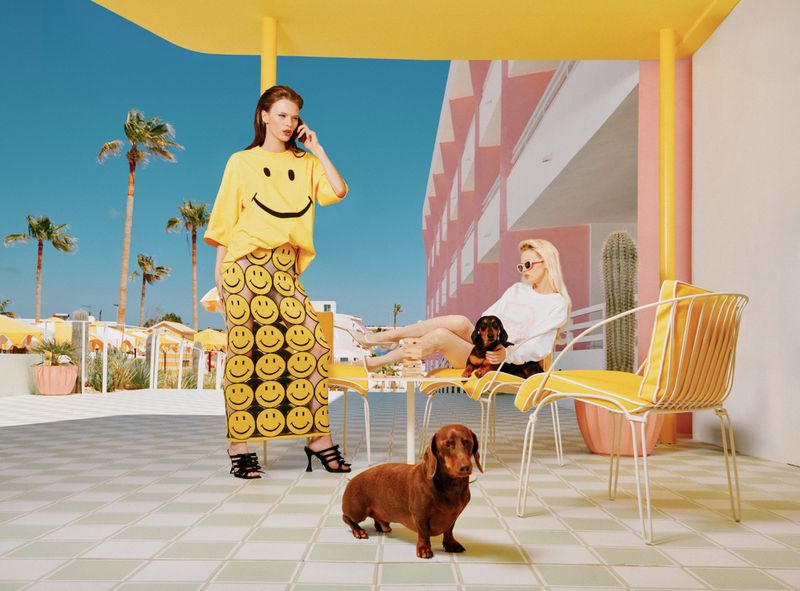 Two female models and a dachshund dog are posing in a vibrant outdoor setting, showcasing yellow, black smiley face apparel and sunny ambiance.