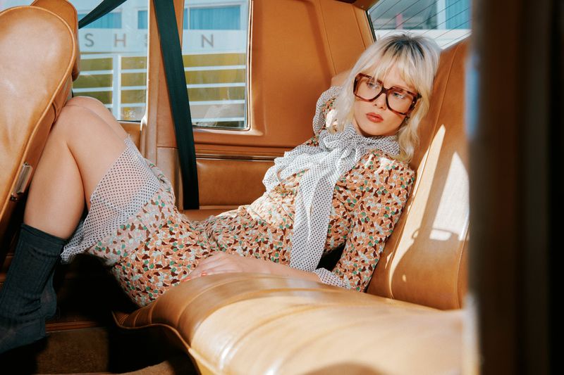 A stylish adult female with glasses and a patterned dress poses inside an old car with tan interior and a building visible outside.