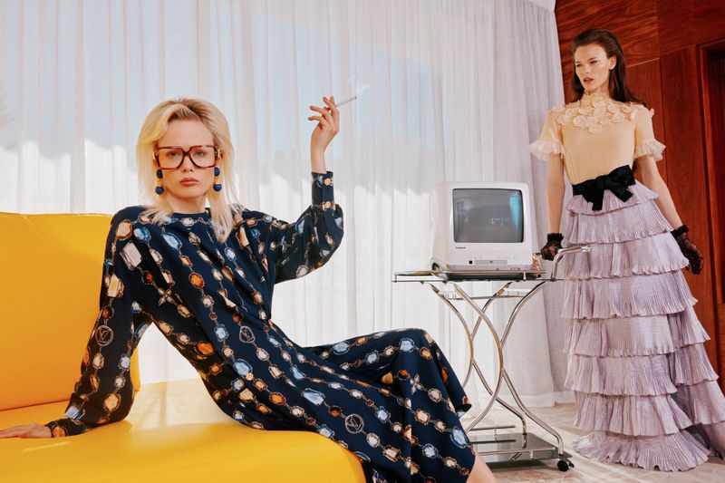 Two fashion models pose in retro-inspired clothing with a vintage television, set against a backdrop of white curtains and warm wood tones.