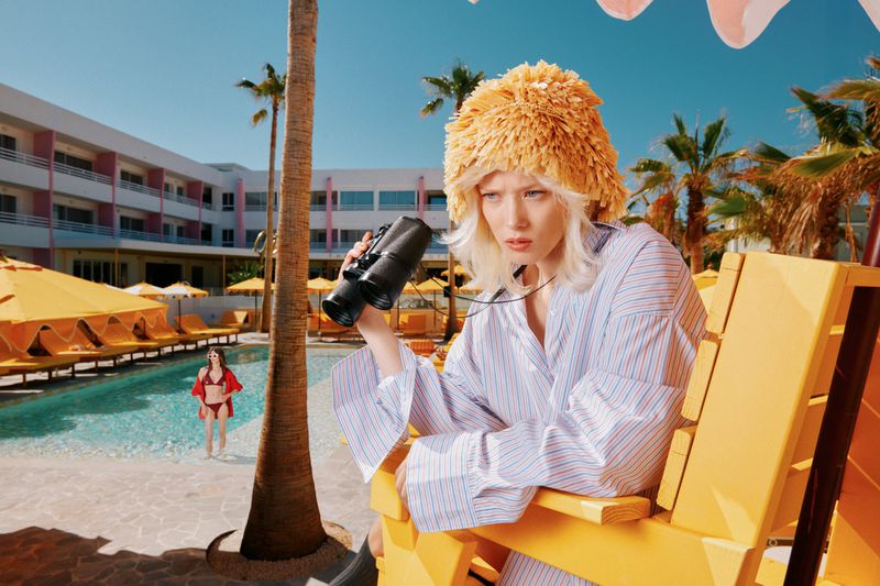 Woman in striped shirt and yellow hat looks through binoculars near a pool with hotel and palm trees under a clear blue sky.