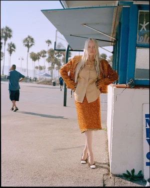 Model in orange tweed jacket and matching skirt standing outdoors near a blue beach kiosk with palm trees in the background