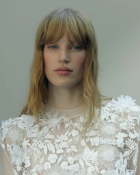 Portrait of a female model with long blonde hair and bangs wearing a sheer white floral embroidered top against a plain light background