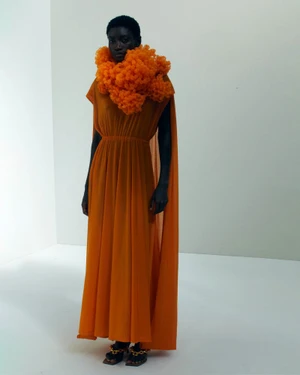 Model wearing a long orange pleated dress and ruffled neck scarf standing against a white seamless background