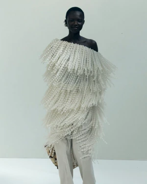 Model wearing a textured off-the-shoulder white fringed top and pale trousers against a white seamless background.