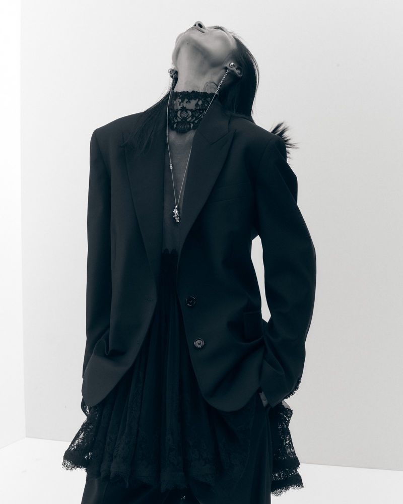 Model in black blazer and lace turtleneck tilting head back against a white seamless background