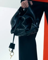Close-up of a person holding a black leather handbag with gathered details and dangling key charms against a white background.