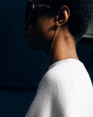 Close-up profile of a model wearing dark sunglasses, gold chain earrings, and a white top against a dark blue background.