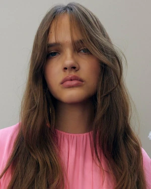 Close up portrait of a young female model with long brown hair wearing a pink top against a neutral light grey background.
