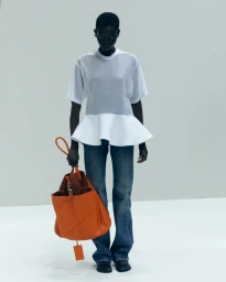 Model standing against a white seamless background wearing a white peplum top, blue jeans, and holding a large orange leather bag.