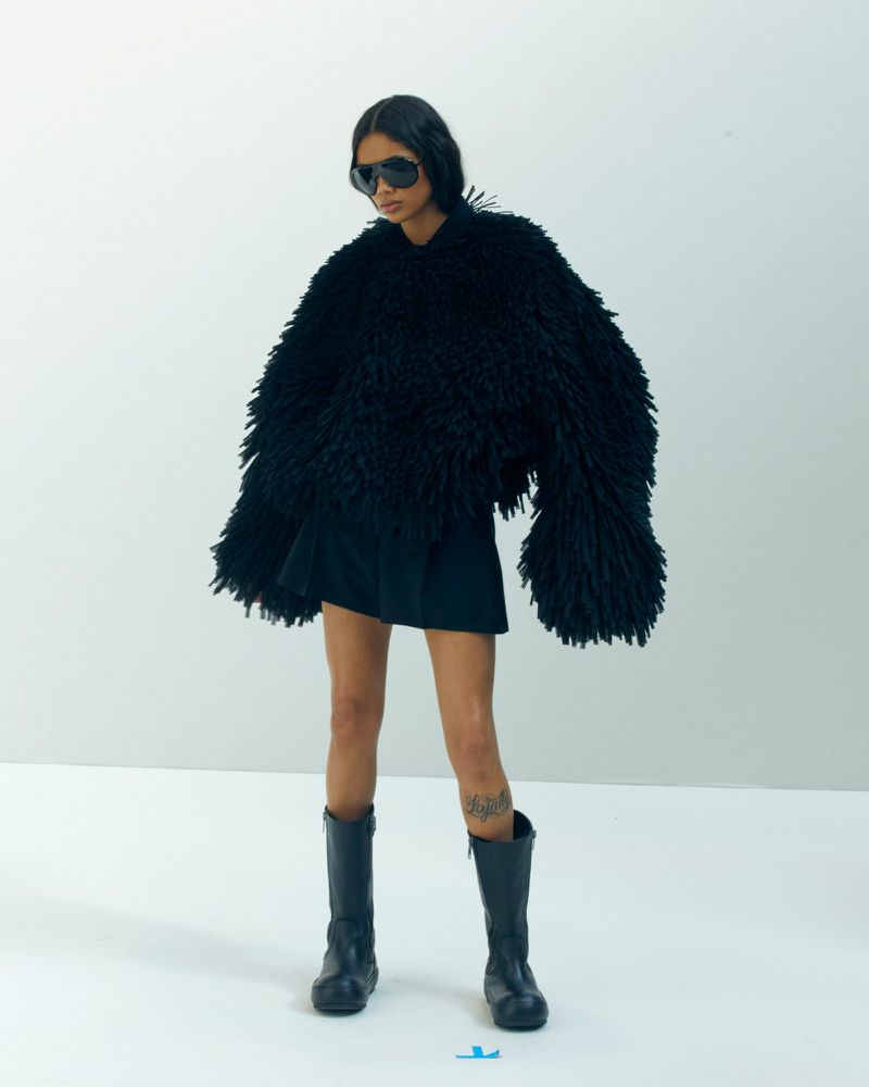 Model in black sunglasses wearing a shaggy textured oversized jacket and tall boots against a white background