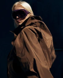 Model wearing dark tinted goggles and a brown hooded coat against a dark background