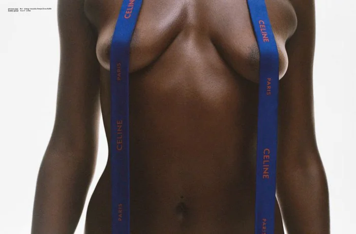 Close-up of a topless Black woman wearing blue branded ribbon straps against a plain white background