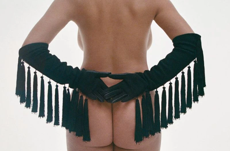Rear view of a nude model wearing long black fringed gloves against a plain white background
