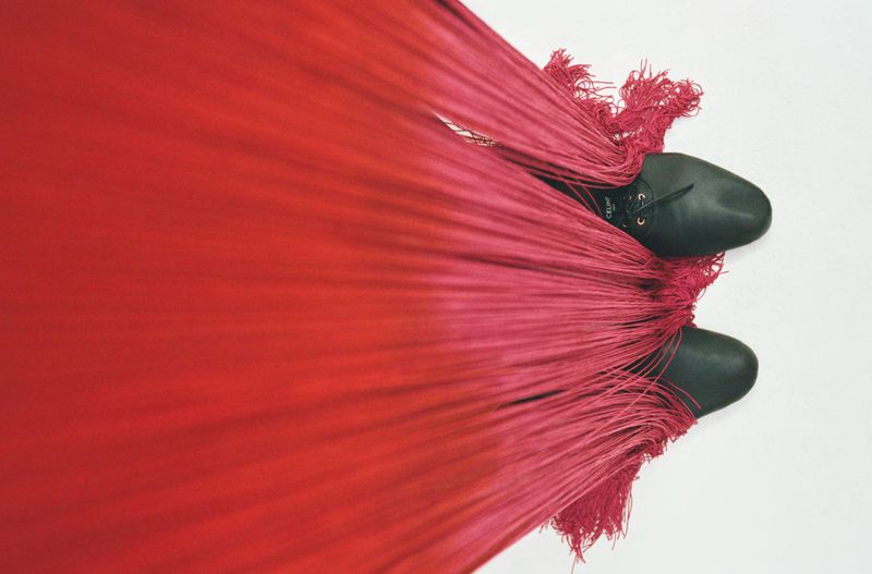 Overhead view of two black shoes peeking out beneath a heavy cascade of long red fringe fabric against a white background.