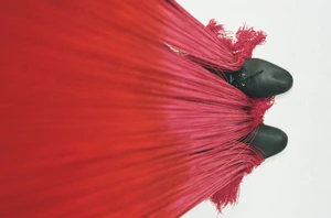 Overhead view of two black shoes peeking out beneath a heavy cascade of long red fringe fabric against a white background.