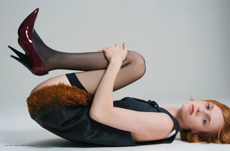 Model lying on her back with legs curled up wearing black heels, sheer tights, and a dark dress against a light background.