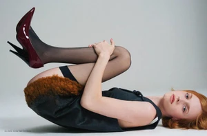 Model lying on her back with legs curled up wearing black heels, sheer tights, and a dark dress against a light background.